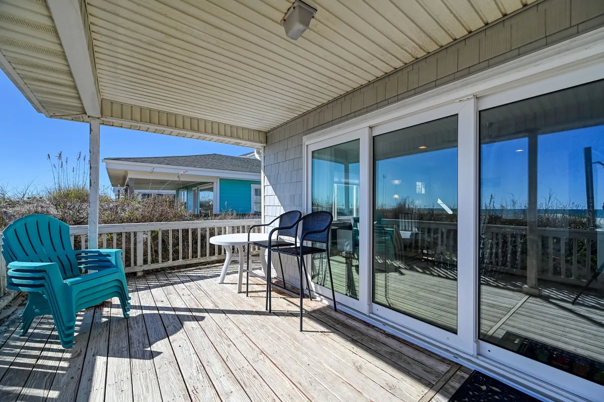 An oceanfront porch or deck, furnished with blue Adirondack chairs and a table, set before sliding glass doors that connect the outdoor space with the interior. The scene suggests relaxed outdoor living, with direct access for enjoying breezes and sunsets over the ocean.