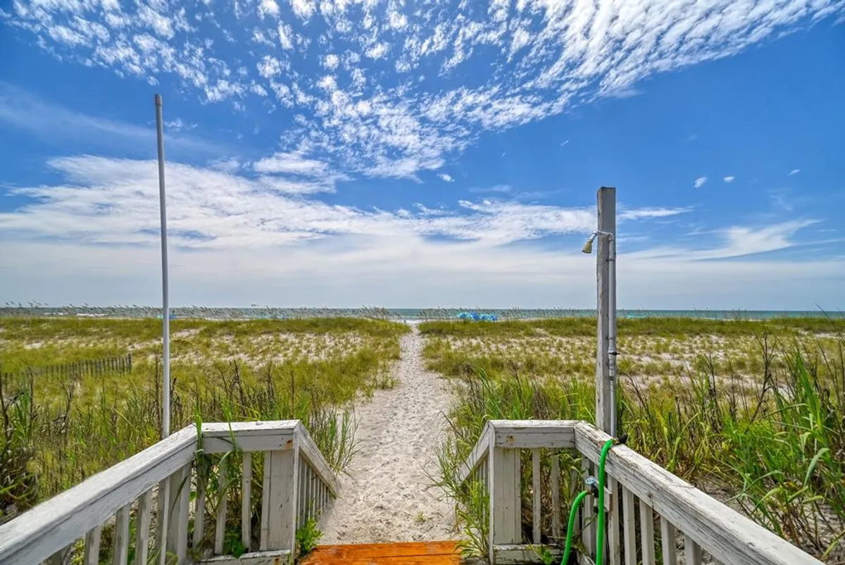 A wooden staircase leads from a private boardwalk down through grassy sand dunes towards the expansive, blue ocean. The path is flanked by dune grass, and the horizon stretches under a serene sky. The view invites immediate access from the home directly onto the beach.