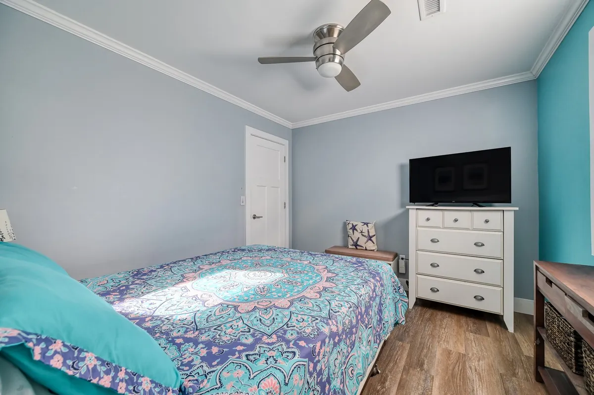 A cozy bedroom featuring a bed with patterned bedding, a television on a dresser, and an accent chair. Natural light spills into the room, creating a tranquil and inviting atmosphere.
