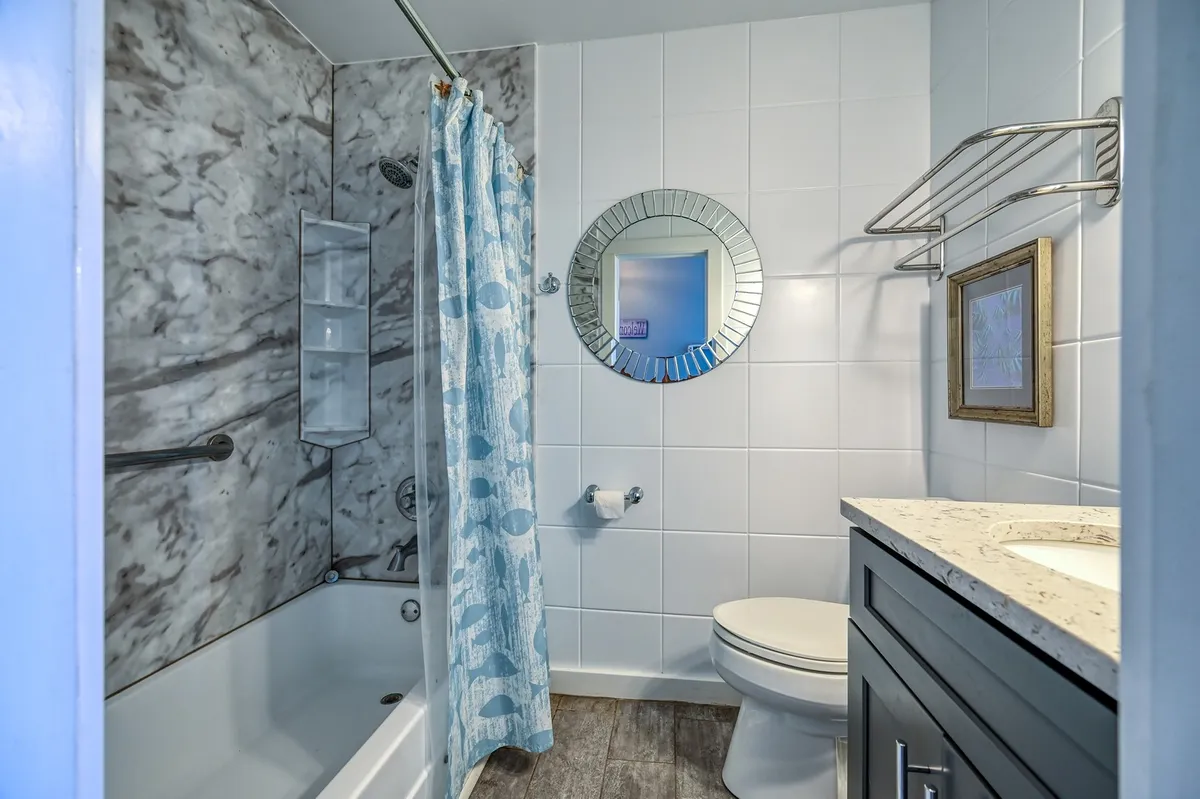 A bright and inviting bathroom featuring a bathtub, mirror, shower curtain, and sink. The space is designed for relaxation, with upgraded fixtures creating a sense of affordable luxury.