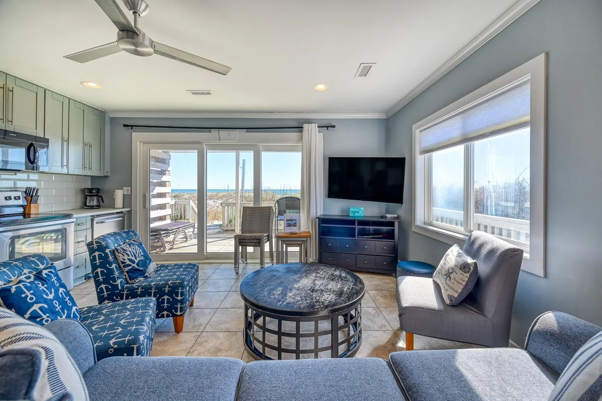 This image showcases a spacious, open-concept living area with comfortable seating, a coffee table, large windows framing the ocean, a kitchen area visible in the background, and a smart television. Natural light floods the space, enhancing the coastal atmosphere and providing a direct connection to the beautiful ocean views outside.
