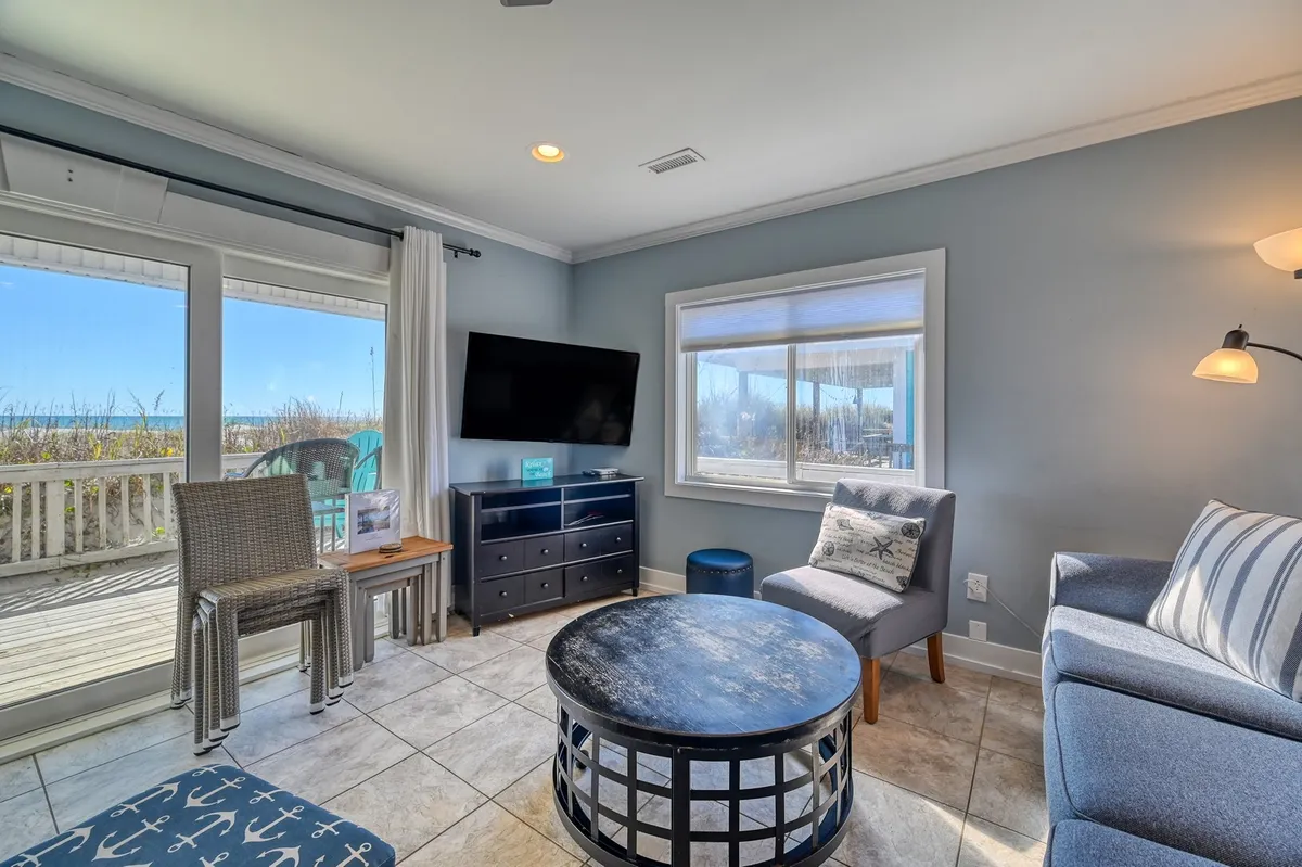 The image showcases a cozy, inviting living space with comfortable seating arranged around a stylish coffee table. Large windows frame picturesque ocean views, filling the room with natural light and offering the perfect spot to relax, watch the sunset, or gather with friends and family after a day at the beach.