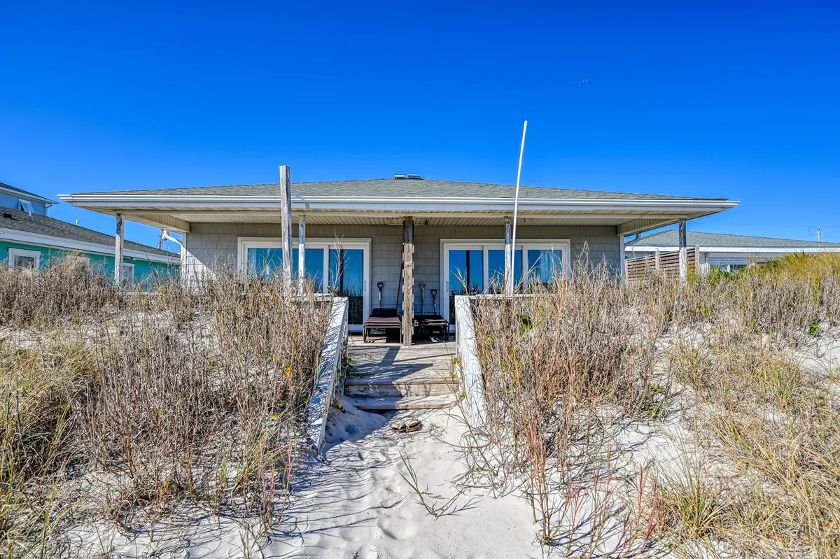 This image showcases the exterior of the beach house, featuring a welcoming wraparound porch, a sandy pathway, and lush surrounding vegetation. The tranquil setting invites guests to relax outdoors or head straight to the nearby beach, making it easy to envision morning walks and time spent in nature with a pet.