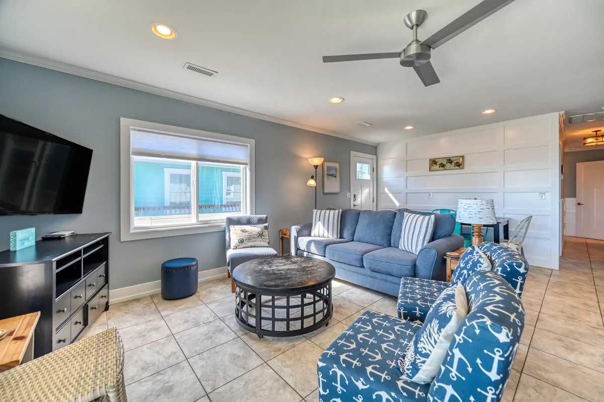 A cozy living space featuring a comfortable blue sofa, anchor-patterned chairs, a circular coffee table, a TV on the wall, and a large window filling the room with sunlight. The view out the window hints at the coastal surroundings.