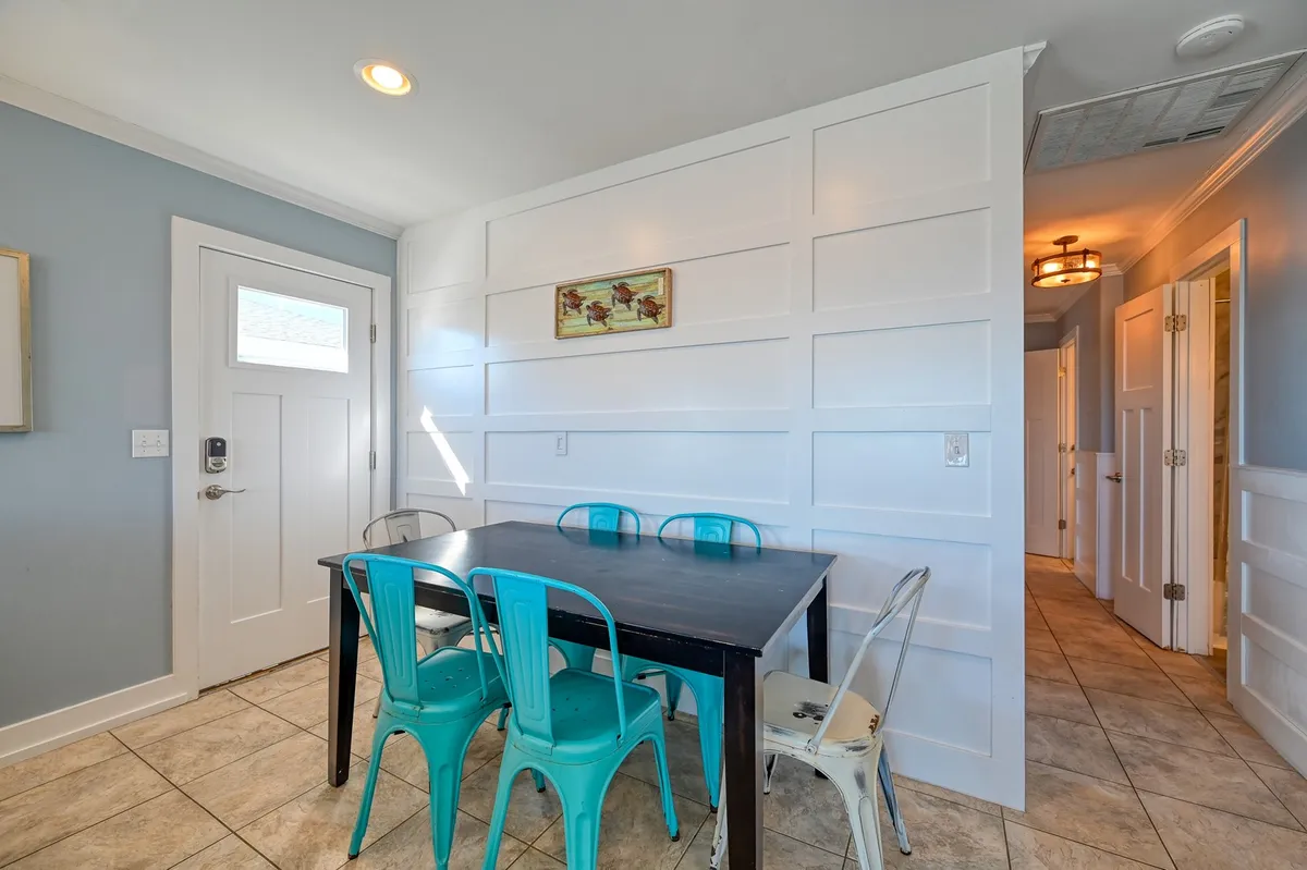 A cozy dining area with a table for six, surrounded by chairs, subtle wall paneling, and a nearby doorway leading to other parts of the home. The setting feels welcoming and perfect for shared meals or planning seaside adventures.