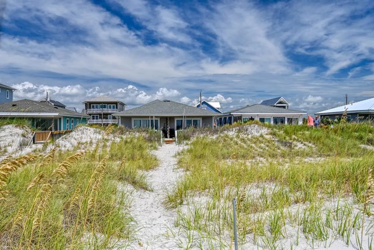 A tranquil scene of beachfront homes, a sandy pathway winding through dune grass, and clouds drifting across the sky. The image highlights the proximity of the beach houses to the ocean and the inviting environment of Surf City.