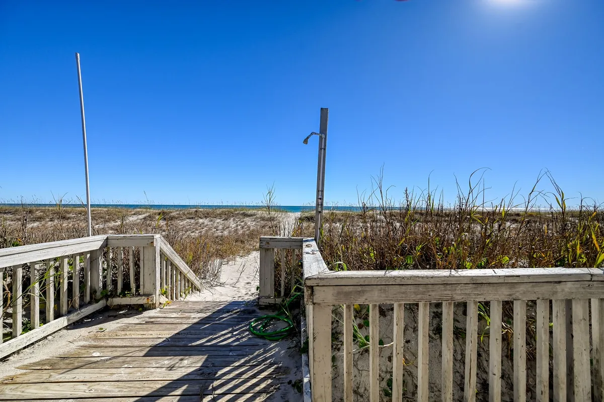 A wooden pathway leads from the porch area over the dunes to the sandy beach. A convenient outdoor shower fixture is visible near the boardwalk, illustrating easy beach access and clean-up after a day in the sand.